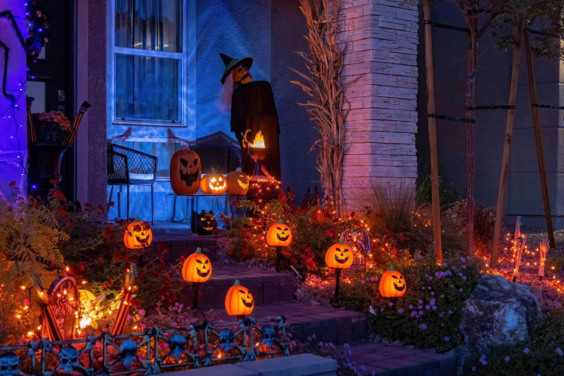 Spooktacular lights A skeleton decoration wearing a witch hat is propped up behind a porch lit up with jack-o-lantern lights at night.